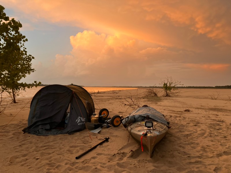 A kayak and a small tent on sand next to a river at sunset