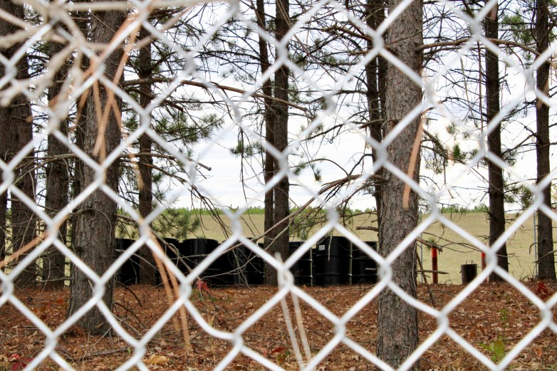 Several black barrels are seen through a chain-link fence in a wooded area with tall trees and fallen leaves and an open green space in the distance.