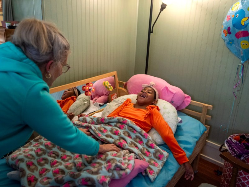 A person wearing a blue sweatshirt leans over another smiling person lying on a pillow in a bed and wearing an orange top, covered with a patterned blanket, with a floor lamp and a colorful balloon beside the bed.
