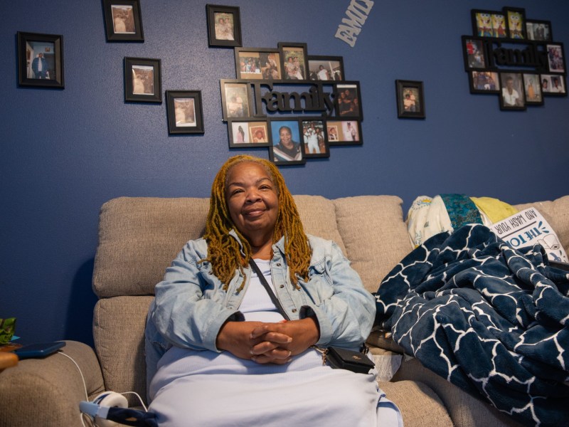 A person sits on a beige couch with hands folded, with blankets on the couch and framed photos and "Family" lettering on a blue wall.