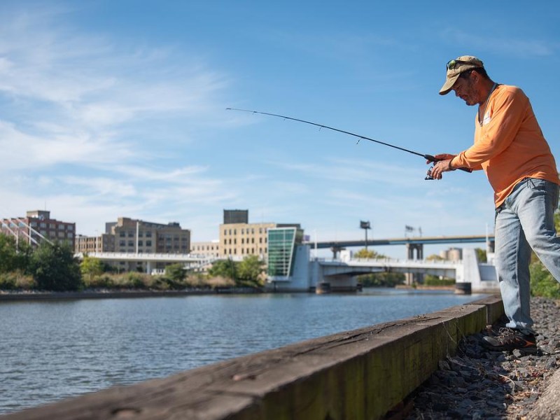 A person wearing an orange shirt and cap fishes from the edge of a riverbank with bridges and buildings visible across the water.