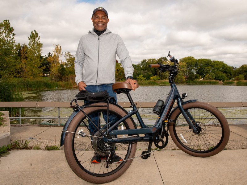 A person wearing a light jacket and cap stands next to a bicycle on a paved path near a body of water with trees in the background.