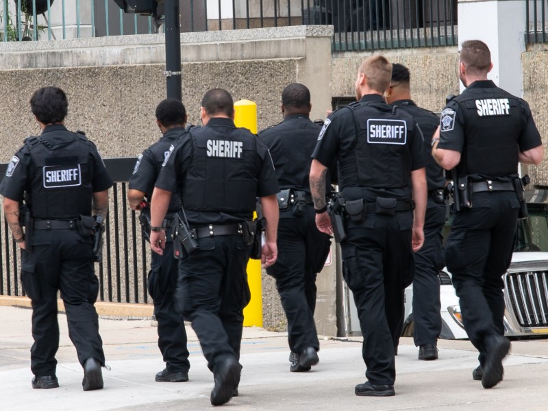 A group of people wearing uniforms labeled "SHERIFF" walk on a sidewalk near a concrete building and a parked vehicle.