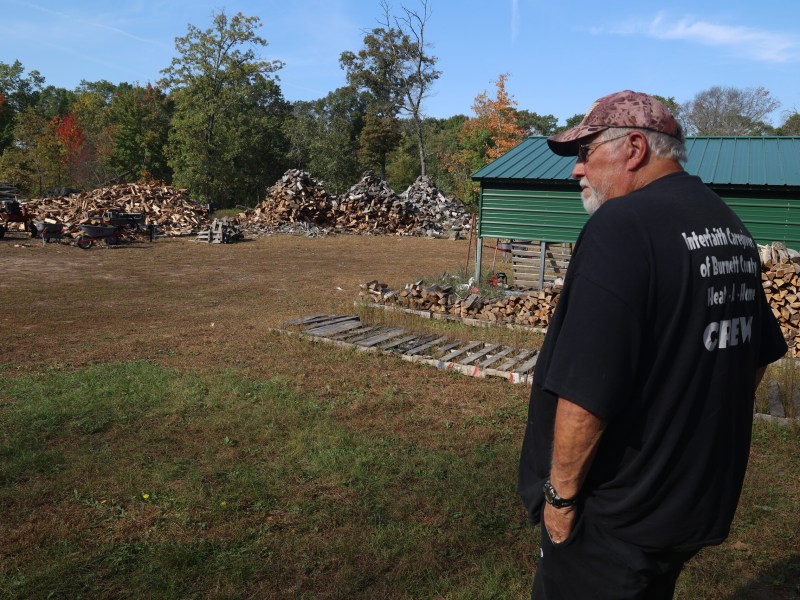 A person, wearing a shirt that reads "Interfaith … Burnett County … Crew," stands near stacked firewood and pallets beside a green shed, looking across a yard with large wood piles and wheelbarrows.