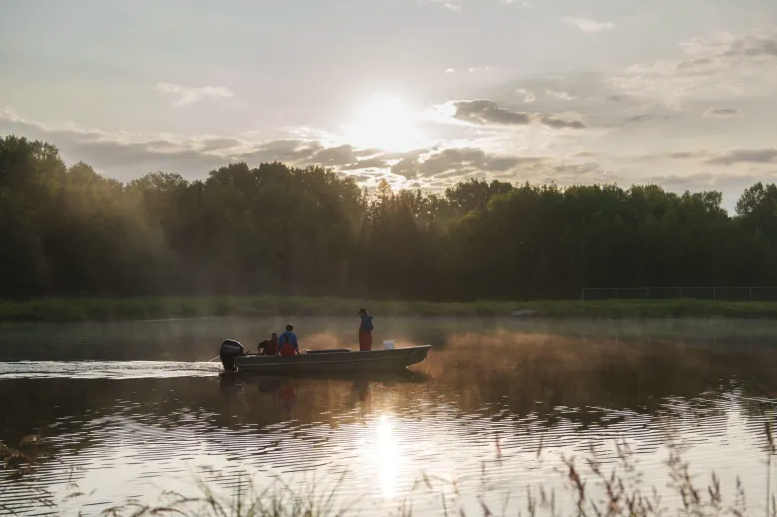 Three people in a small motorboat on water with mist rising and the sun shining through trees in the background.