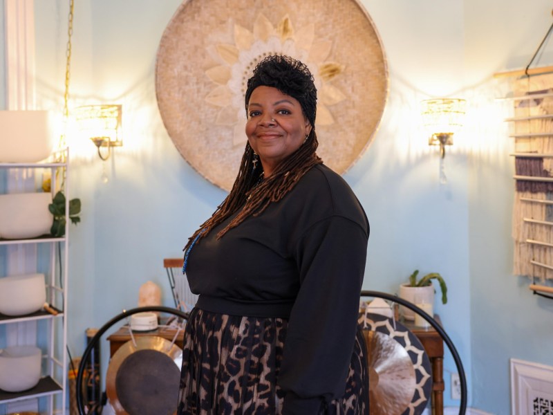A person stands indoors smiling toward the camera, wearing a dark top and patterned skirt, with art on light blue walls, lamps, shelves, and decorative objects visible behind the person.