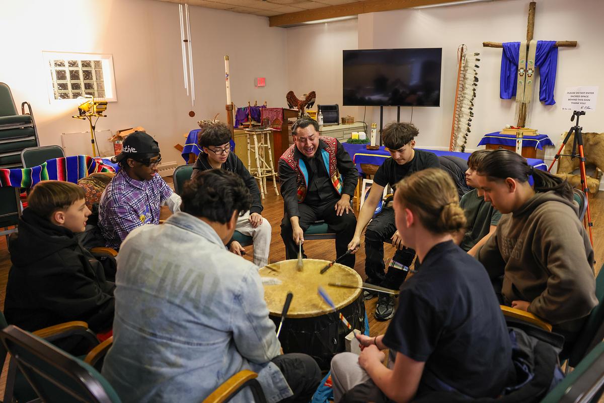 People sit in a circle striking a large drum with padded sticks inside a room with chairs, blankets and ceremonial items displayed on the walls.