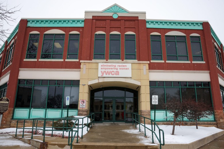 A two-story brick building with green trim displays a sign reading “eliminating racism empowering women YWCA” above the entrance, with light snow on the ground in front.