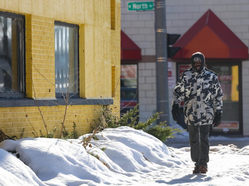 A person walks along a snowy sidewalk past a yellow brick building, wearing a hooded patterned jacket and gloves, with a street sign reading "North Ave" in the background.