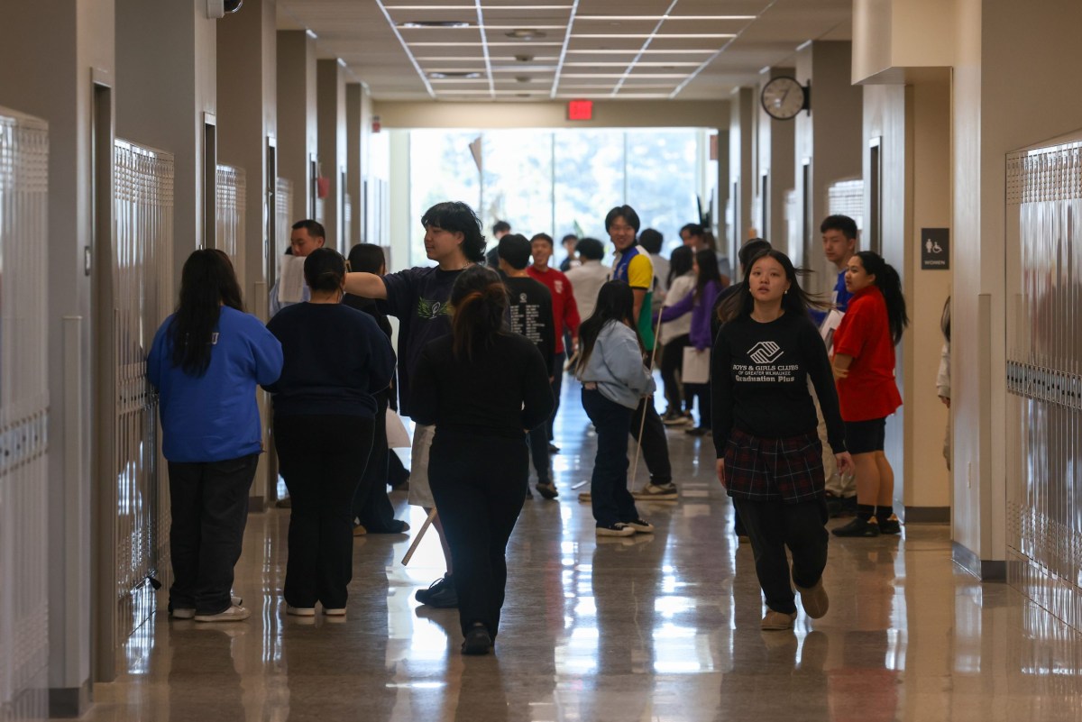 People stand and walk in a hallway lined with lockers, including a person wearing a shirt reading "Boys & Girls Clubs of Greater Milwaukee," with a "Women" restroom sign on a wall and a red "EXIT" sign above a window at the end of the hallway.