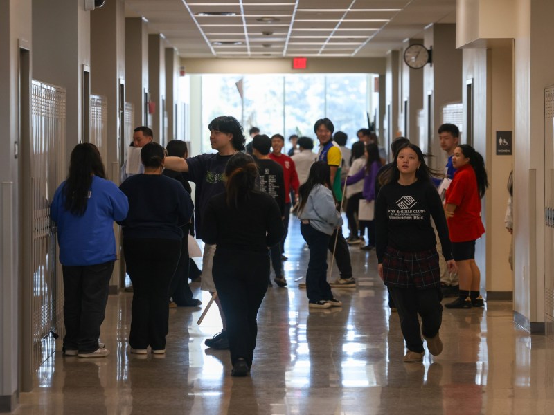 People stand and walk in a hallway lined with lockers, including a person wearing a shirt reading "Boys & Girls Clubs of Greater Milwaukee," with a "Women" restroom sign on a wall and a red "EXIT" sign above a window at the end of the hallway.