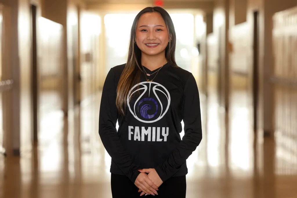 A person stands in a hallway lined with lockers, wearing a shirt with a basketball graphic and the word "FAMILY" printed below it.