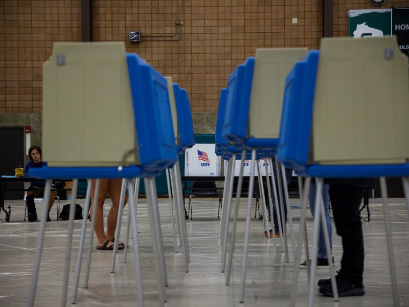 People stand at blue voting booths in a large indoor space as a person sits at a table in the background near signs reading "VOTE."