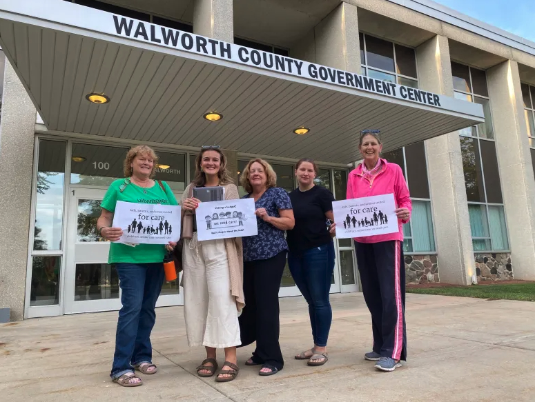 Five people stand holding signs reading “for care” outside a building labeled “WALWORTH COUNTY GOVERNMENT CENTER” near entrance doors.