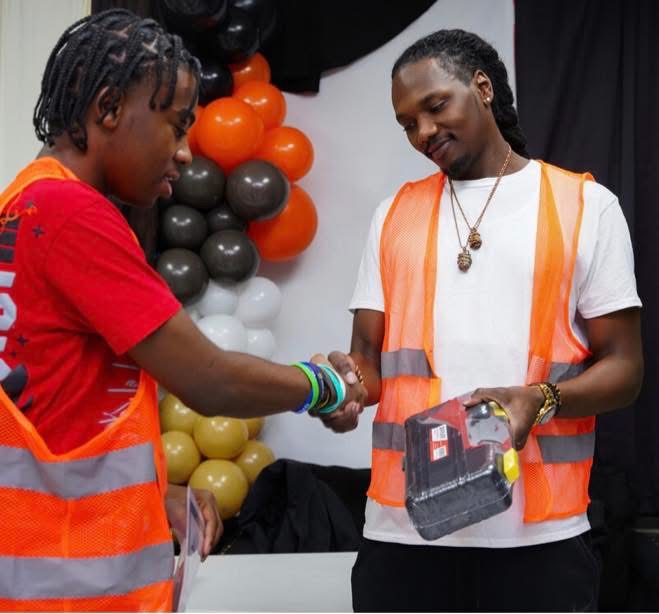Two people wearing orange safety vests shake hands, with one person holding a small boxed item, and black, orange, white and gold balloons in the background