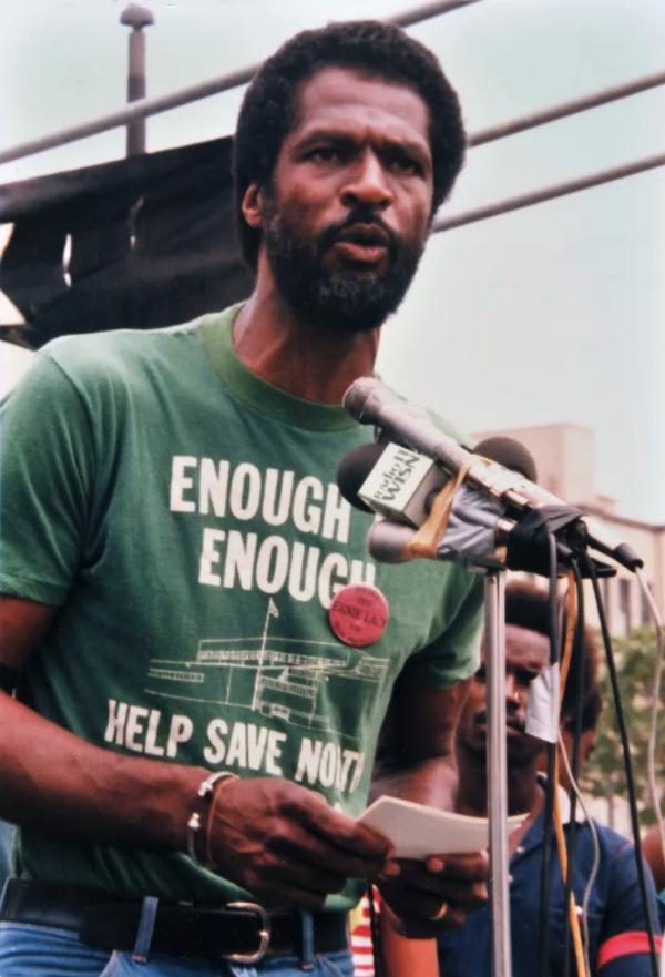 A person speaks into multiple microphones while holding papers, wearing a green shirt reading "ENOUGH IS ENOUGH HELP SAVE NORTH," with others standing in the background.