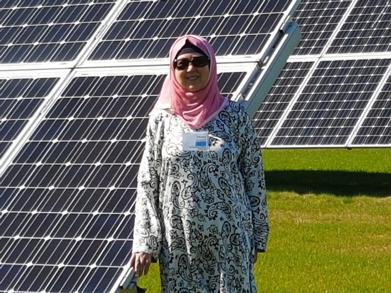 A person wearing sunglasses and a pink headscarf stands on grass in front of rows of tilted solar panels in bright sunlight.