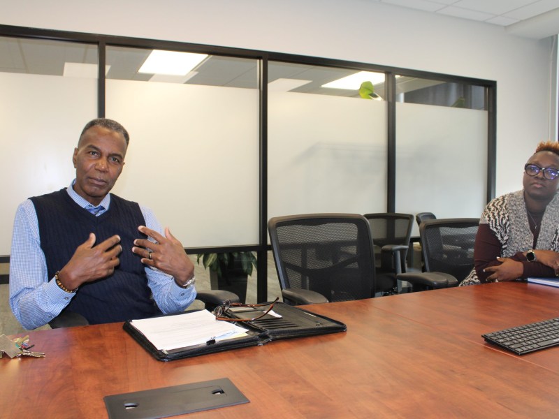 Two people sit at a wooden conference table in a room with glass walls, one person gesturing with hands, and papers, keys and a keyboard on the table.