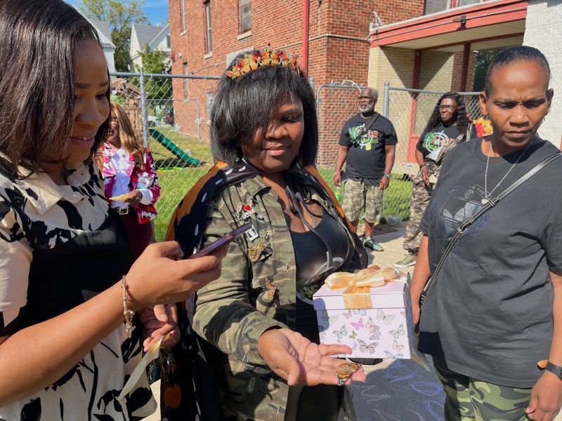 A person wearing a camouflage jacket holds a butterfly in one hand next to a pink gift box with butterfly images while standing outside a brick building with five other people nearby.