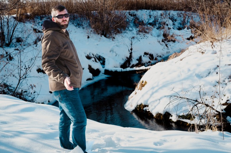 A person wearing sunglasses, a brown coat and blue jeans stands in snow beside a narrow creek with snow-covered banks and leafless shrubs, gesturing with one hand while facing the camera.