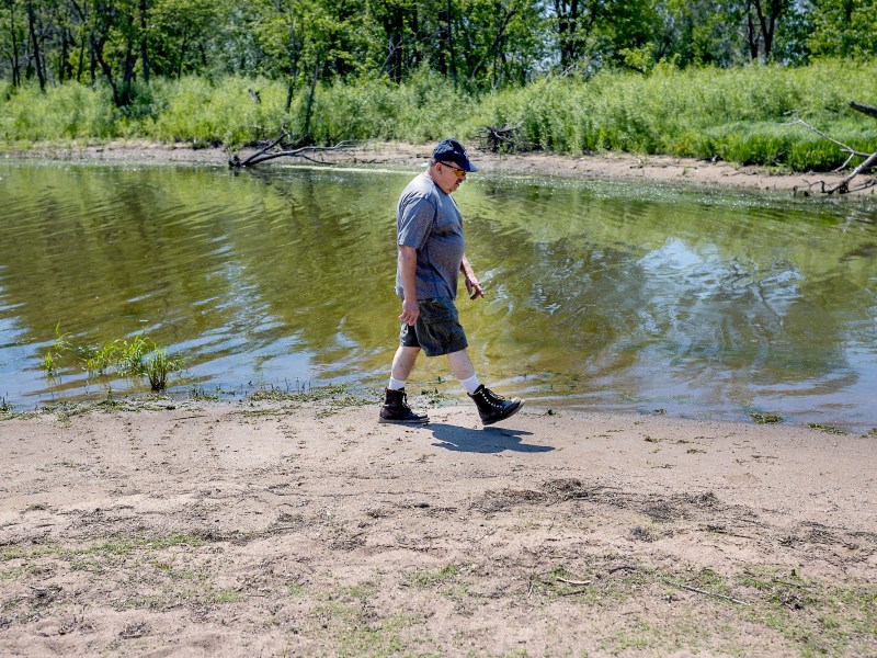 A person wearing a hat, T-shirt, shorts, and boots walks along a sandy bank beside calm water with green trees and grass in the background.