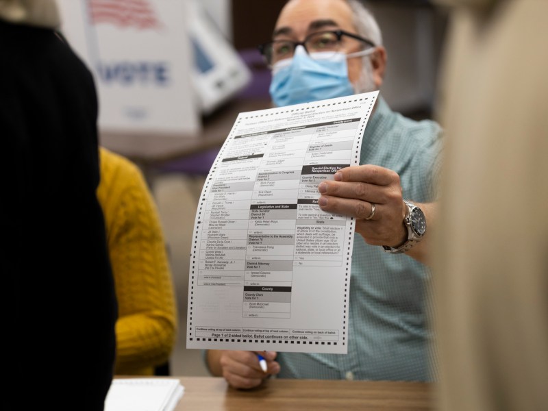 A person wearing a face mask holds up a paper ballot with printed candidate lists while seated at a table, with other people partially visible nearby.