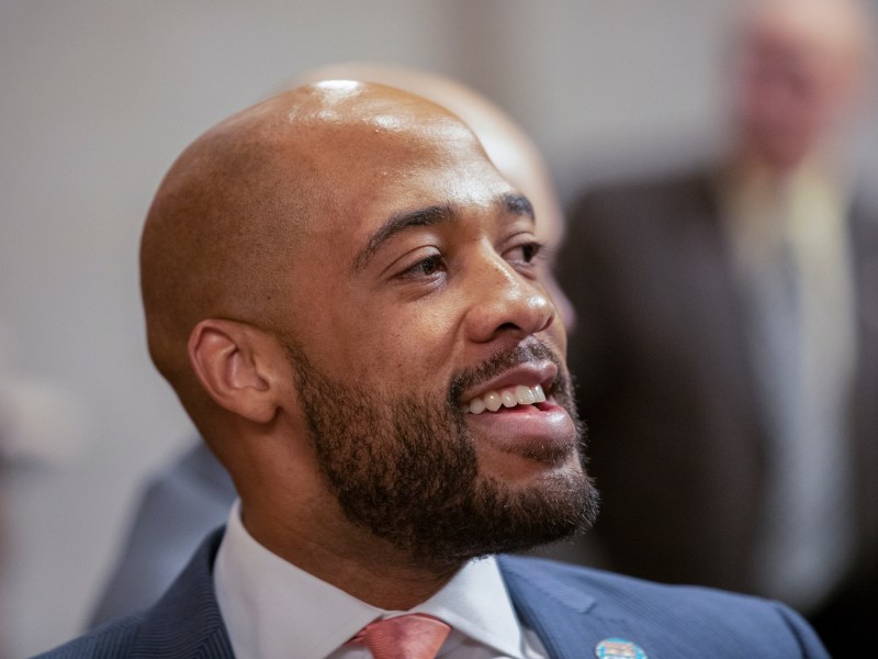 A person in a blue suit and reddish tie looks to the side while smiling, with blurred people in the background.