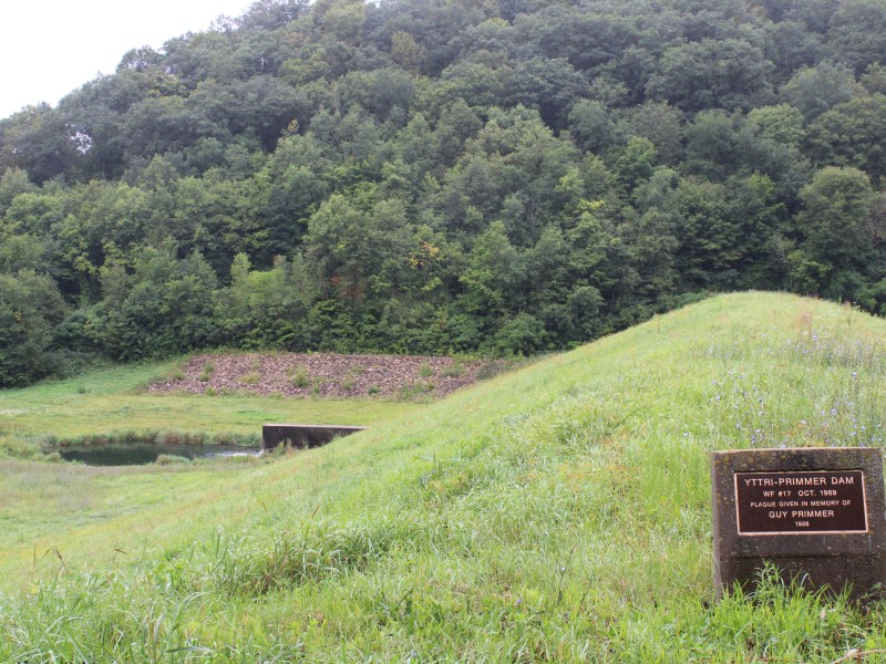 A plaque on a stone base says “YTTRI-PRIMMER DAM” in the foreground with a grassy hill, water and trees behind it.