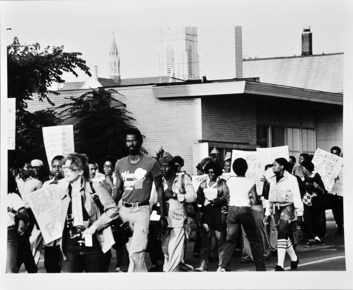 People walk on a street holding signs, including one reading "EQUAL RIGHTS," with buildings and a church steeple in the background.