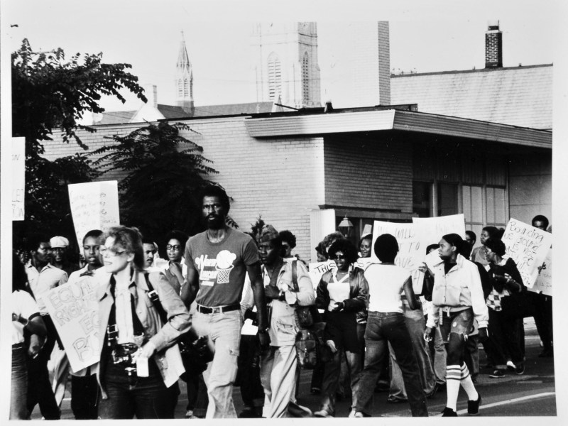 People walk on a street holding signs, including one reading "EQUAL RIGHTS," with buildings and a church steeple in the background.