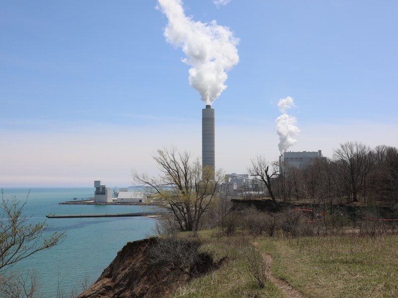 Two smoke plumes billow into a blue sky at a power plant next to a lake.