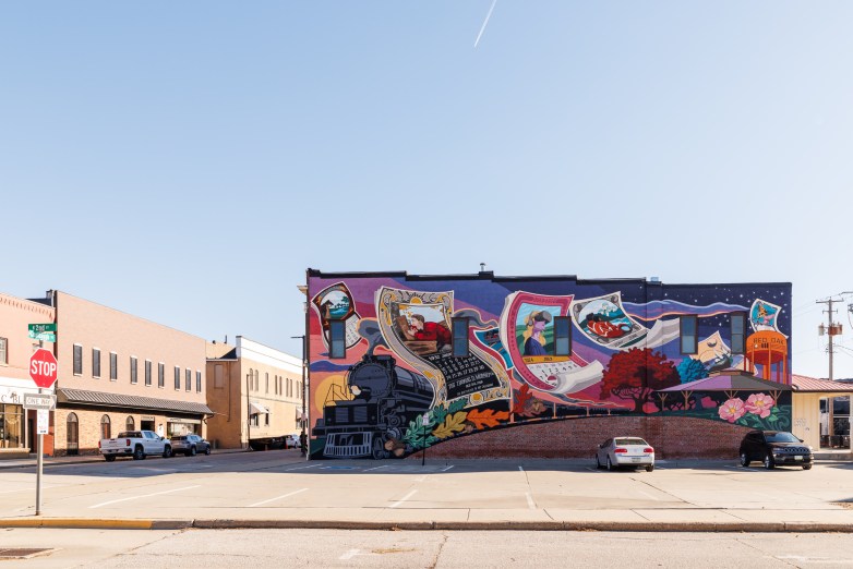 A colorful mural covers the side of a building, depicting a train, calendar pages and an orange water tower labeled "RED OAK," with parked cars in front and on a street and other buildings nearby.
