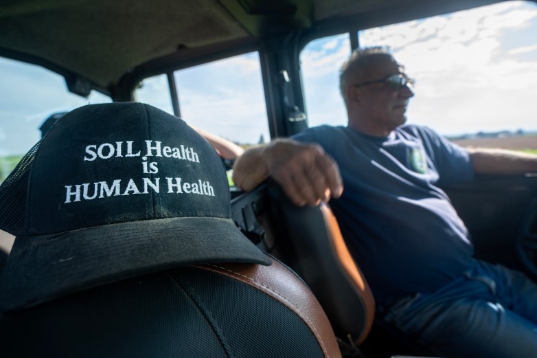 A black cap on a vehicle seat reads "SOIL Health is HUMAN Health," with a person sitting in the driver’s seat looking to the right with an out-of-focus field in the background.