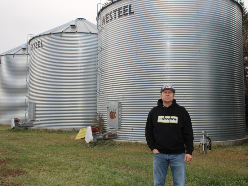 A person wearing a hooded sweatshirt with "STEIGER" on the front stands on grass in front of several large corrugated metal grain bins with "WESTEEL" on them.