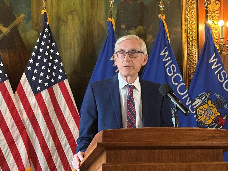 A person stands at a wooden podium with a microphone, flanked by U.S. flags and blue flags reading “Wisconsin” inside an ornate room.