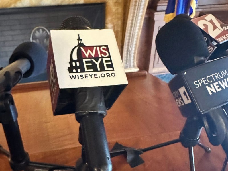 Several microphones on stands are next to a wooden podium, with logos on the microphones reading “WIS EYE,” “SPECTRUM NEWS 1” and “27 WKOW”